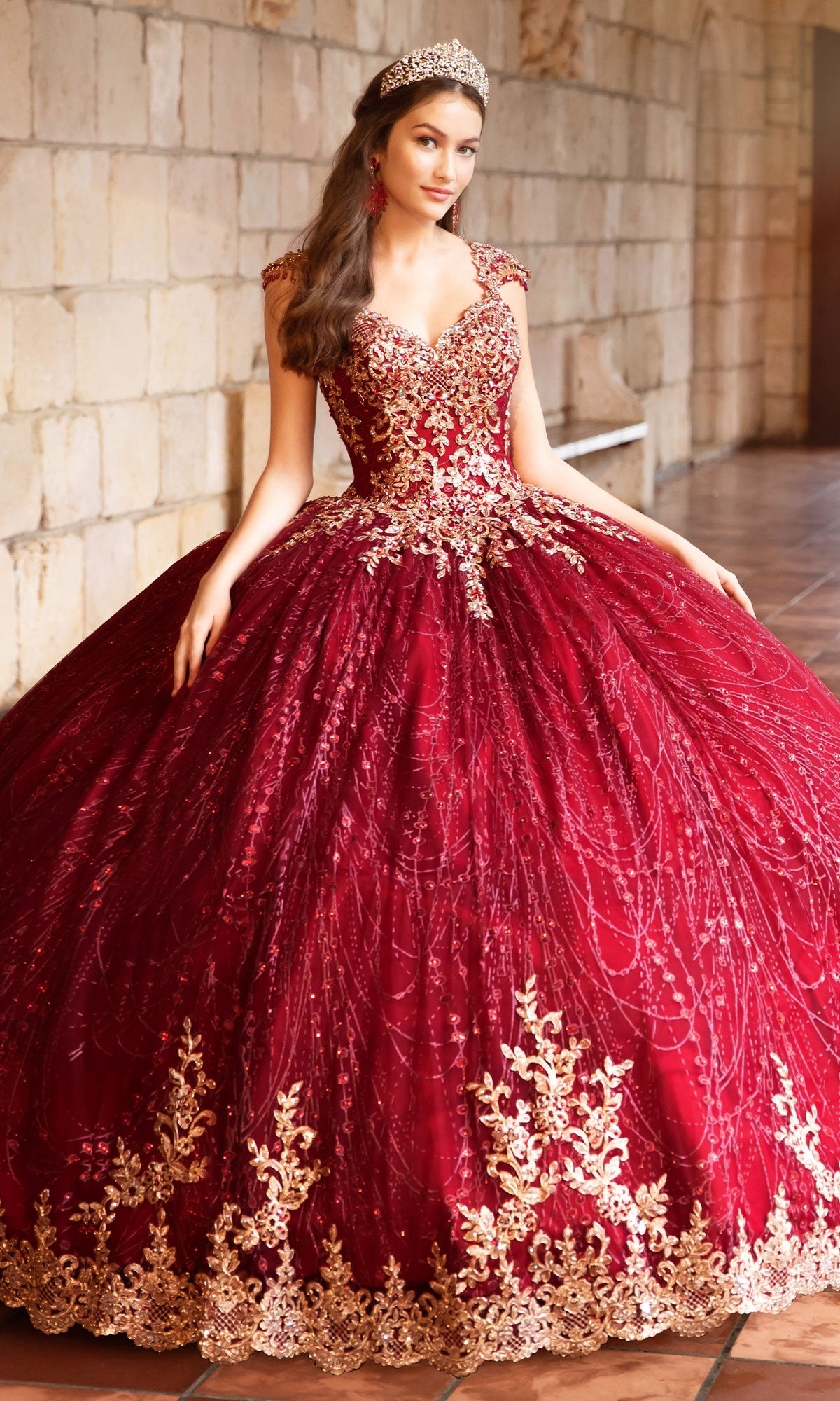 A young woman in the Princesa PR21964 Cap-Sleeve Quinceanera Dress, featuring gold embroidery and a corset bodice, with a tiara, stands in a stone hallway.